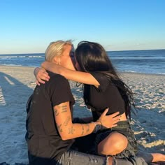 two people sitting on the beach kissing each other and one person is holding his arm around the other's neck