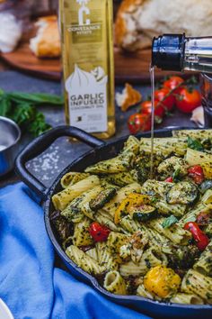 a skillet filled with pasta and vegetables being poured over the top by a bottle of olive oil