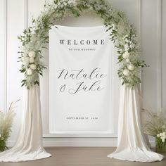 a welcome sign with flowers and greenery on the front is displayed in front of two wedding archs