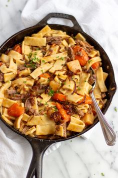 a skillet filled with pasta and meat on top of a white tablecloth next to a spoon