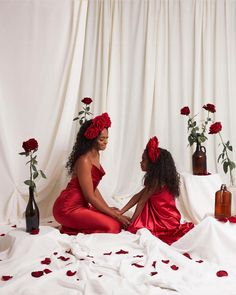 two women in red dresses sitting on a bed with rose petals and roses around them