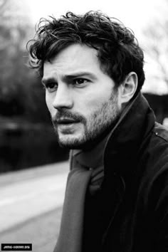 a black and white photo of a man with curly hair wearing a jacket looking at the camera