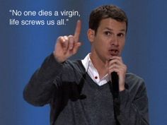 a man holding a microphone making the peace sign with his hand and saying no one dies a virgin, life screws us all