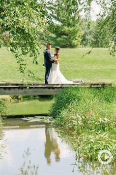 a bride and groom standing on a bridge over a pond in a park with green grass