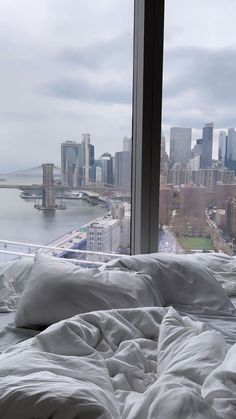 an unmade bed in front of a window overlooking a cityscape and bridge