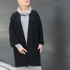 an older woman standing in front of a gray wall wearing a black coat and grey leggings