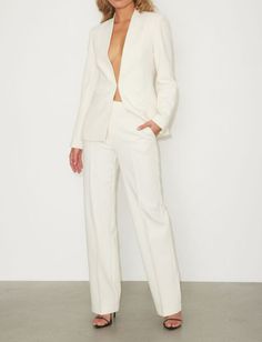 a woman standing in front of a white wall wearing a white suit and black heels