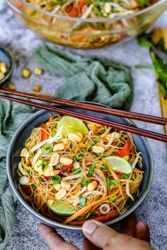 a person holding a bowl of noodles with chopsticks in front of it and another bowl full of food