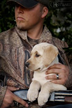 a man holding a white puppy in his lap while wearing a hunting hat and jacket