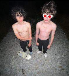 two shirtless boys with skateboards standing in the dirt at night, one wearing red sunglasses