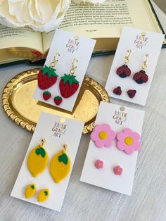 four pairs of strawberry earrings on top of a table with an open book in the background