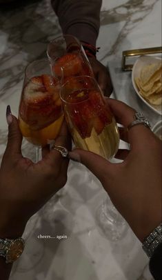 two people toasting with fruit in wine glasses on a marble counter top, while another person holds up a glass to the camera