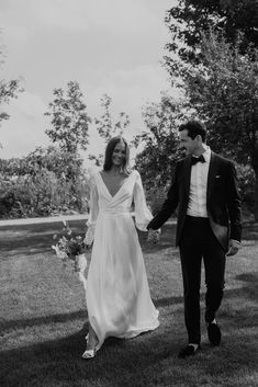 a bride and groom holding hands walking through the grass in black and white photo with trees behind them