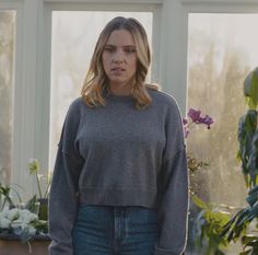 a woman standing in front of a window wearing a gray sweater and jeans with her hands on her hips