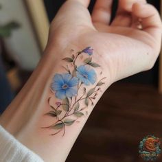 a small blue flower tattoo on the wrist and hand is shown in front of a woman's arm