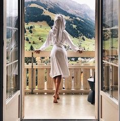 a woman in a robe is standing on a balcony looking out the window at mountains