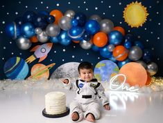 a baby boy in an astronaut suit sitting next to a cake and space themed backdrop