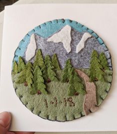 a hand holding a piece of felt with trees and mountains in the background on a white card