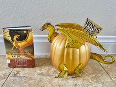 a golden dragon pumpkin sitting on top of a tile floor next to a paperback book