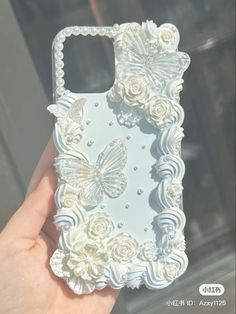 a hand holding an iphone case with white flowers and butterflies on the back, in front of a window