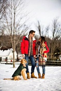 a man and woman are standing in the snow with two dogs, one is wearing a red jacket