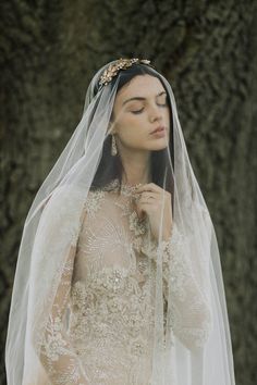 a woman in a wedding dress with a veil on her head is standing near a tree