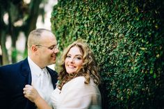 a man and woman standing next to each other in front of a green hedge wall