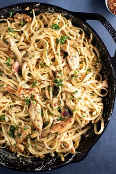 a skillet filled with pasta and chicken on top of a blue tablecloth next to a bowl of seasoning