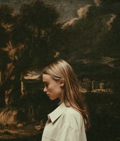 a woman standing in front of a painting with her head turned to the side, wearing a trench coat