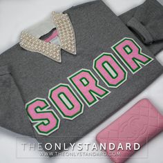 a grey sweatshirt with the word soror on it next to a pink purse and wallet