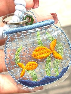 a hand holding a clear plastic case with embroidered fish on it and two white beads hanging from the bottom