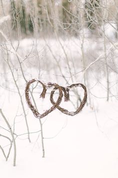 two hearts made out of twigs hanging from a tree in the snow with trees behind them