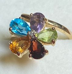 A precious 10K yellow gold semiprecious gemstone flower ring for your consideration. This beauty is hallmarked 10K along with the maker's mark CJC. The pear shaped gemstones are garnet, citrine, peridot, amethyst, and blue topaz. The 6 prong set stones form a colorful flower with a small diamond chip in the center. The gemstones measure approximately 5.5mm x4mm with the setting measuring approximately 12.5mm in diameter. A portion of the setting is actually the bypass portion of the setting for Mexican Fire Opal Ring, Gold Chevron, Fire Opal Ring, Floral Jewellery, Maker's Mark, Size 10 Rings, Multi Stone Ring, Multi Stone, Flower Ring