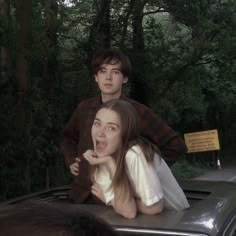 two people sitting on the hood of a car in front of some trees and bushes