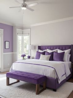 a bedroom with purple and white bedding, pillows, and a bench in front of the window