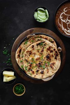 three pita breads on a wooden plate next to bowls of sauce and sliced cucumbers