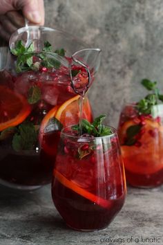 two glasses filled with red liquid and garnished with green leaves, orange slices and mint