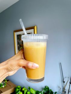 a hand holding a plastic cup with a straw in it next to a plant and pictures on the wall