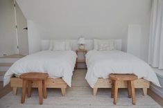 two beds with white linens and wooden foot stools in a room that is painted white