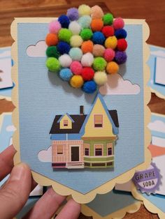 a hand holding up a card with a house and colorful balloons on top of it