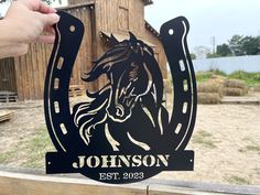 a person holding up a horse sign in front of a wooden fence with a barn behind it