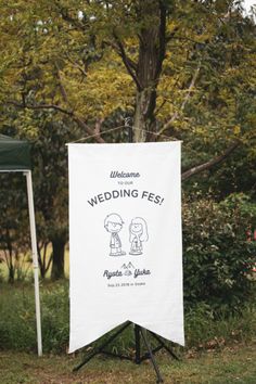 a white sign sitting on top of a grass covered field next to a forest filled with trees