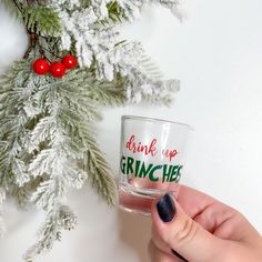 a person holding a glass with the words drink up grinches on it next to a christmas tree