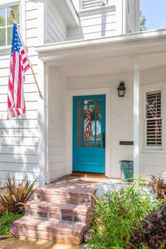 an american flag hanging on the front door of a white house with blue doors and brick steps