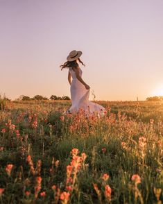 a woman in a white dress and hat walking through a field with wildflowers