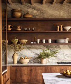 a kitchen with marble counter tops and wooden shelves filled with bowls, plates, cups and vases