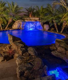 a large pool surrounded by rocks and palm trees at night with blue lights in the water