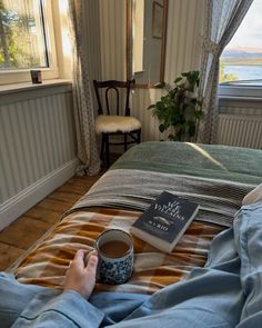 a person laying in bed with a book and cup of coffee on their lap, next to a window