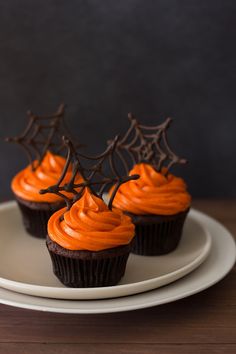 chocolate cupcakes with orange frosting and spider web decorations on top, sitting on a white plate