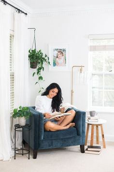 a woman sitting on a blue chair reading a book in her living room with potted plants